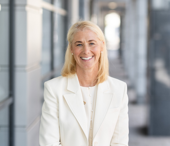 Professional head-and-shoulders portrait of a Jill Britton wearing a white blazer and light-coloured top, standing in front of a modern office building. The background is softly blurred with grey and white architectural elements.